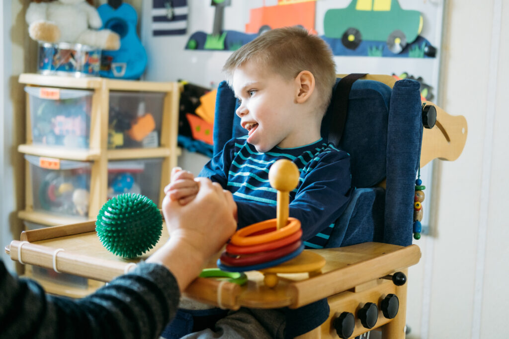 Enfant à besoins particuliers jouant en position adaptée sur une chaise haute avec le soutien d’un parent.
