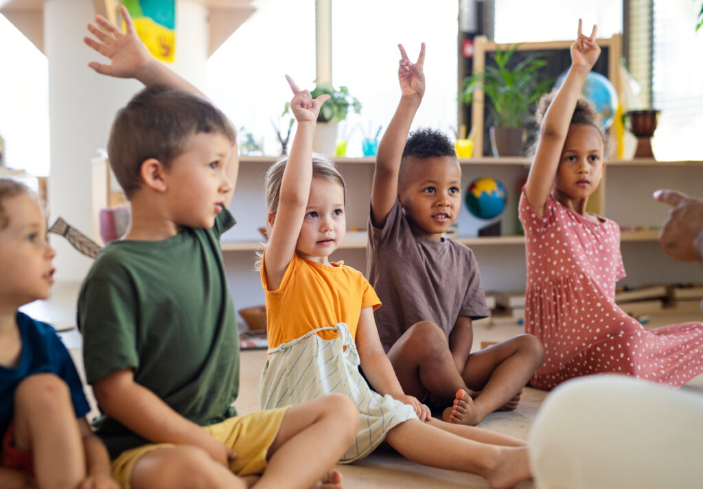 Groupe d’enfants en classe de maternelle, assis au sol et levant la main, favorisant l’apprentissage, la participation et la transition scolaire vers la maternelle.